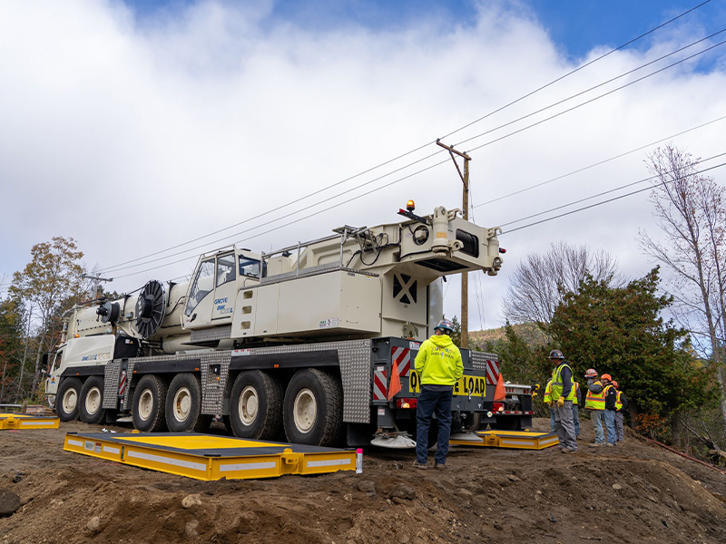 Crane with deployed outriggers; workers nearby under cloudy skies.