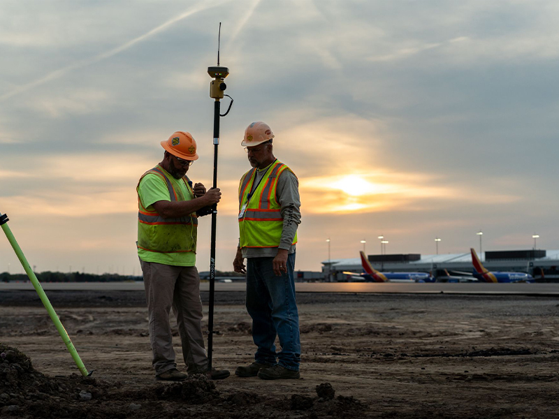Two construction workers survey a dirt site near airport terminal.