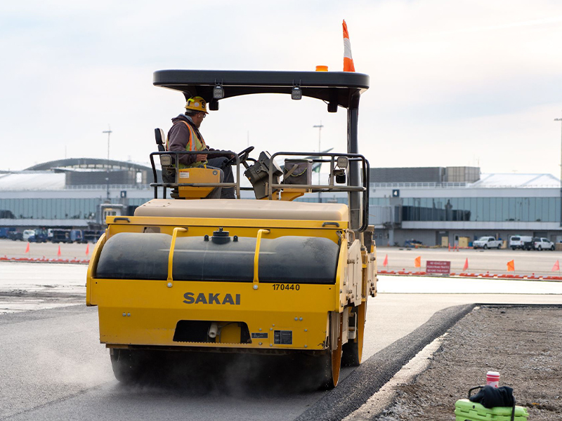 Worker drives yellow steamroller near airport terminal building.