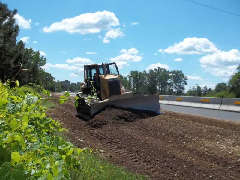 Bulldozer spreads dirt by road under partly cloudy sky.