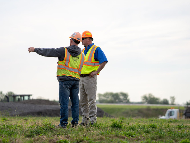 Two workers in safety gear stand on field, with one gesturing ahead