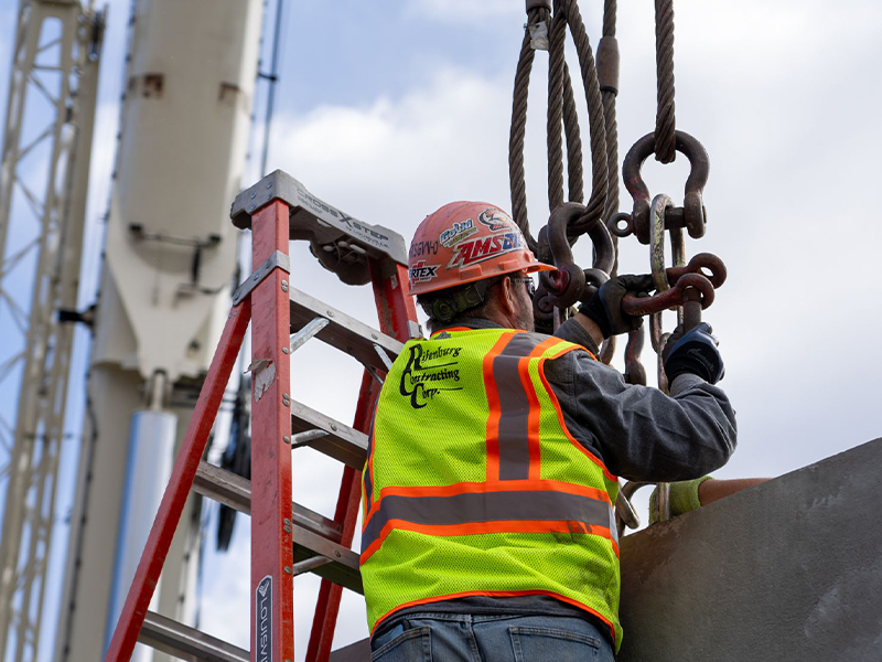 Worker in vest on ladder adjusts metal hooks on cables.