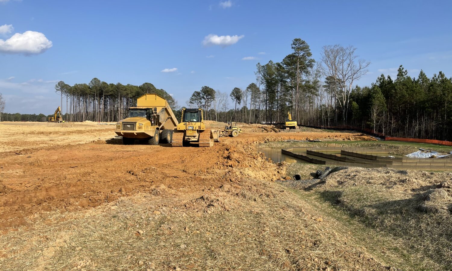 Construction vehicles work near soil piles and pond under blue sky.