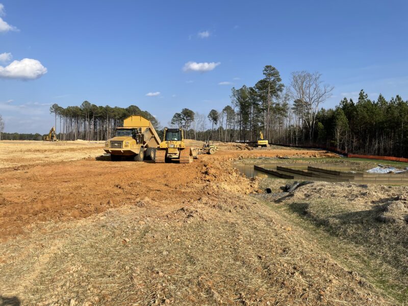 Construction vehicles work near soil piles and pond under blue sky.