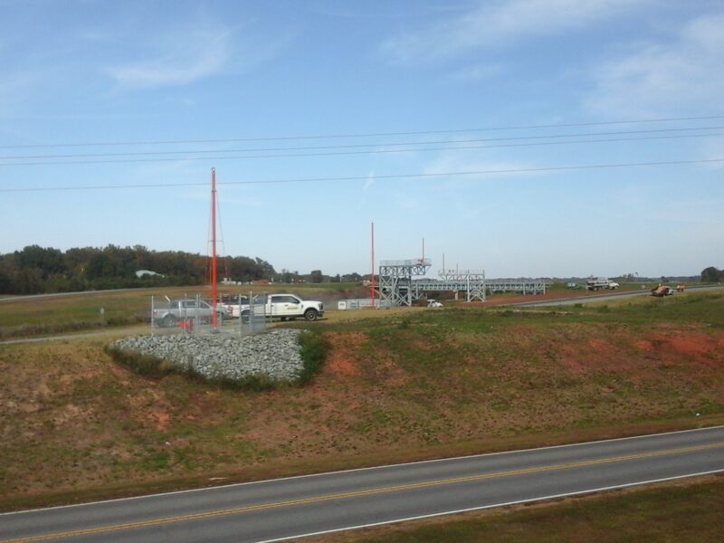 White truck by fenced utility area, fields and equipment behind.