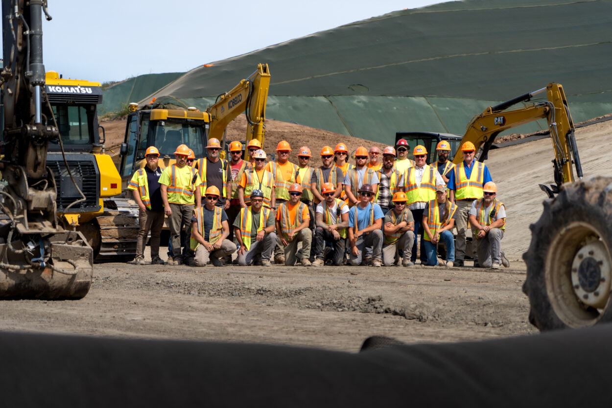 Construction workers in safety gear pose at a site with machinery.