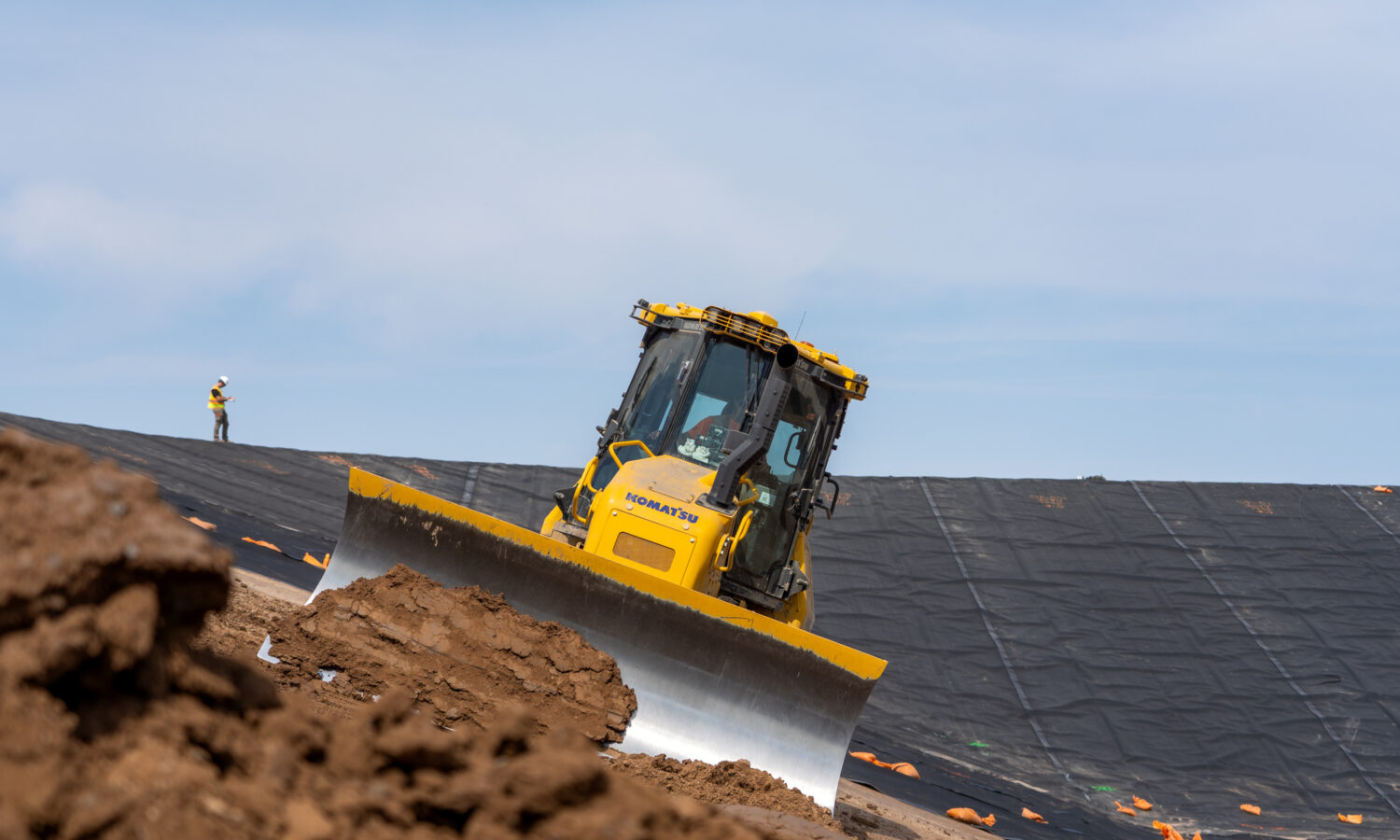 Yellow bulldozer moves dirt; worker stands by tarp-covered slope.