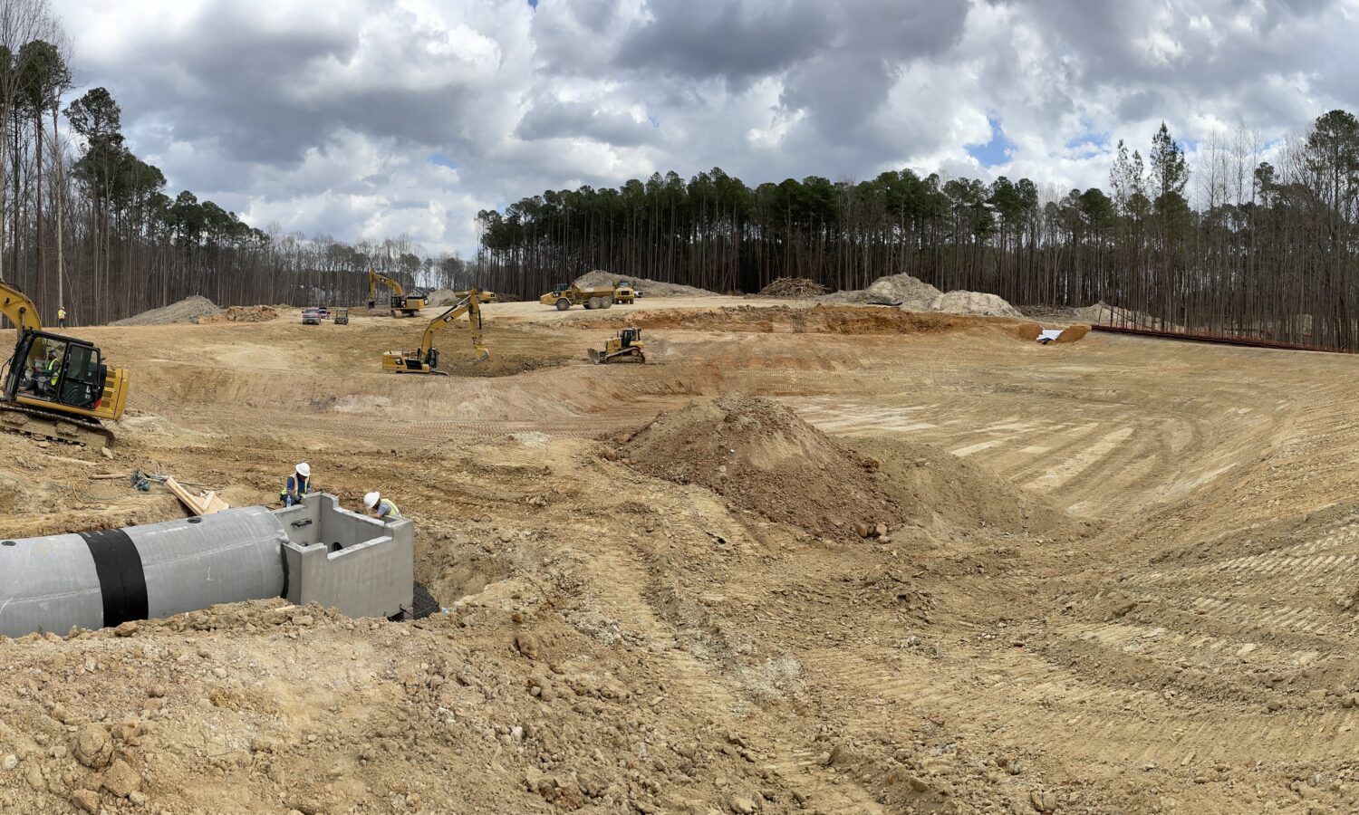 Excavators and worker at construction site; dirt, pipes, trees.