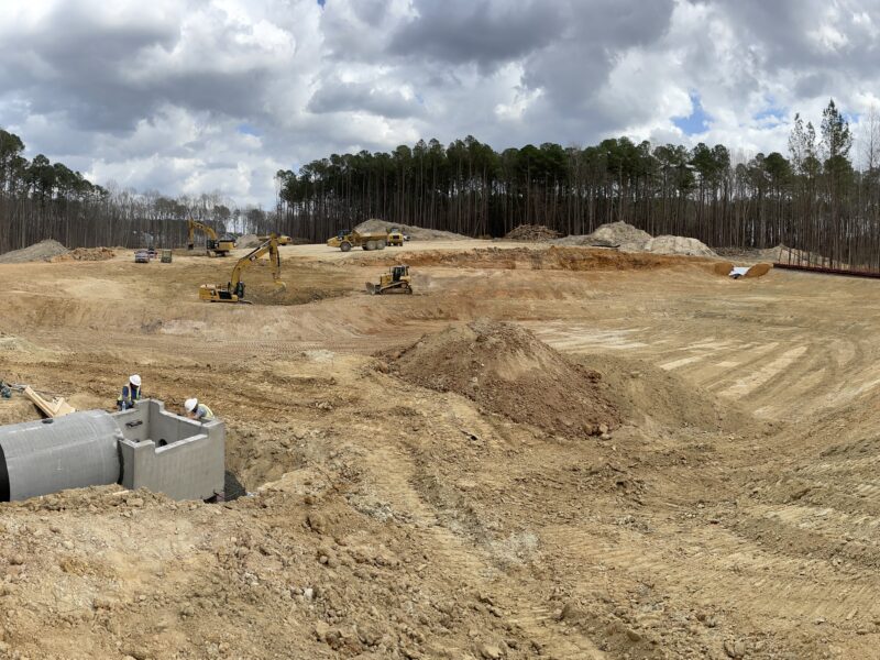 Excavators and worker at construction site; dirt, pipes, trees.
