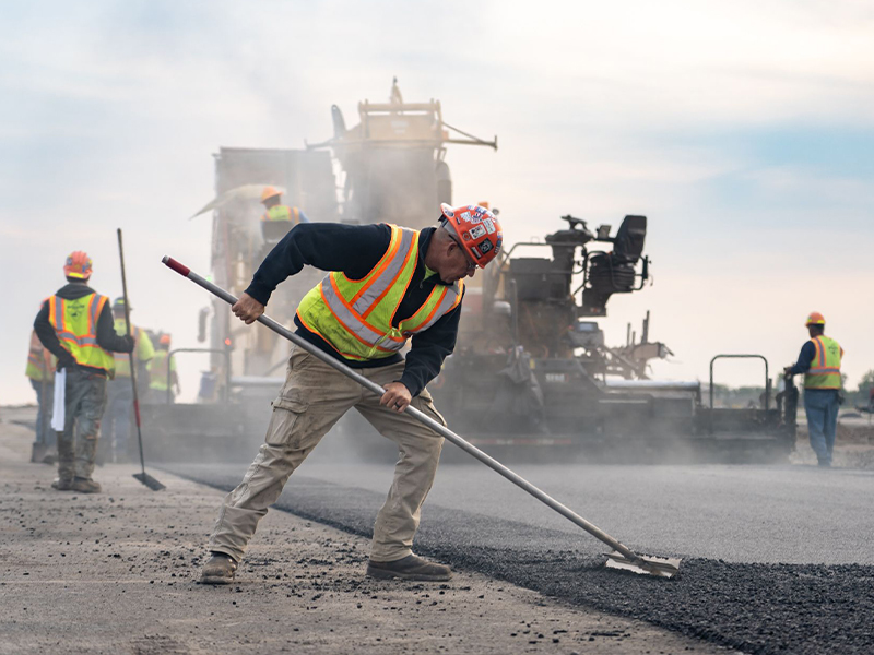 Worker in vest rakes asphalt as others and machines work.