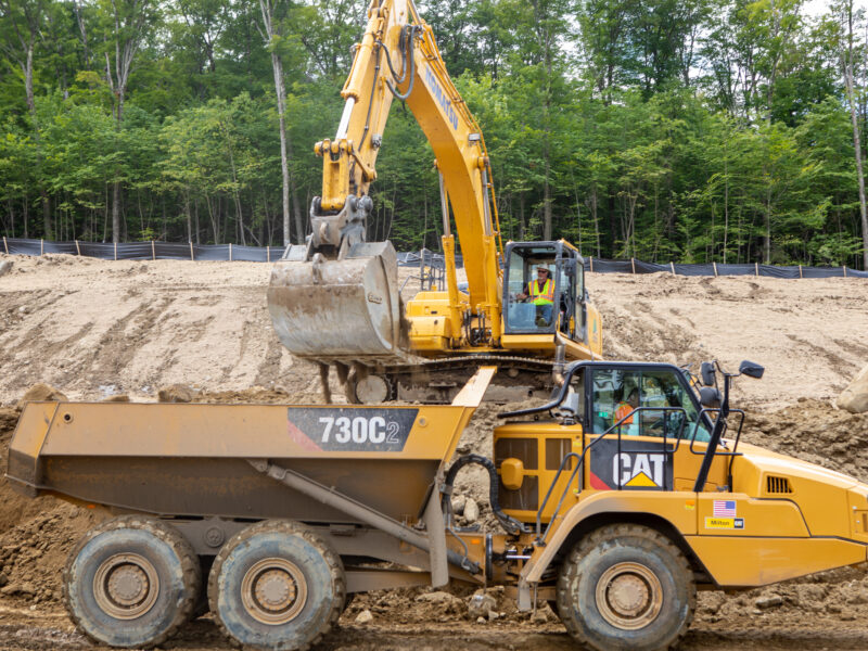 Excavator loads dirt into yellow dump truck at worksite.