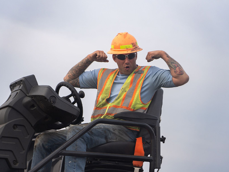 Construction worker flexes arms on equipment in safety gear.