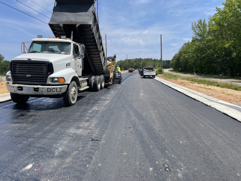Dump truck unloads asphalt; construction vehicles work under clouds.