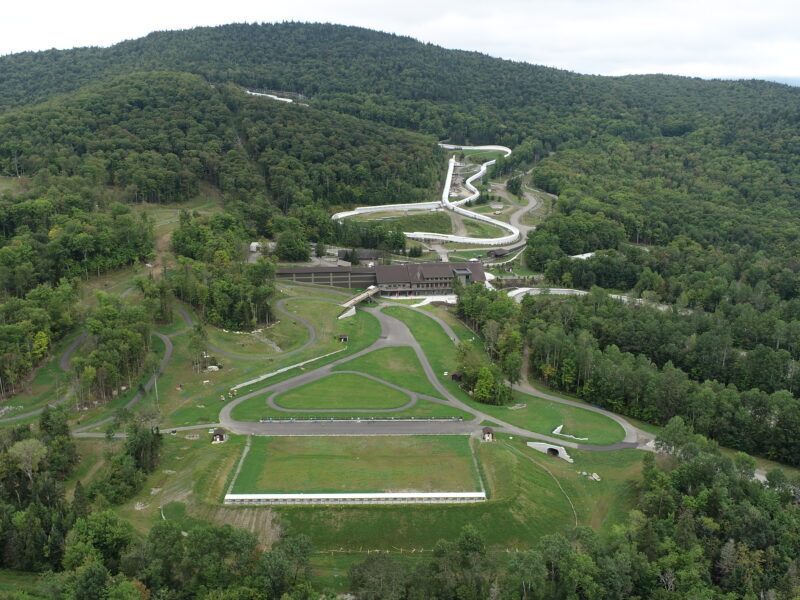 Aerial view: bobsled track winding through dense green forest.
