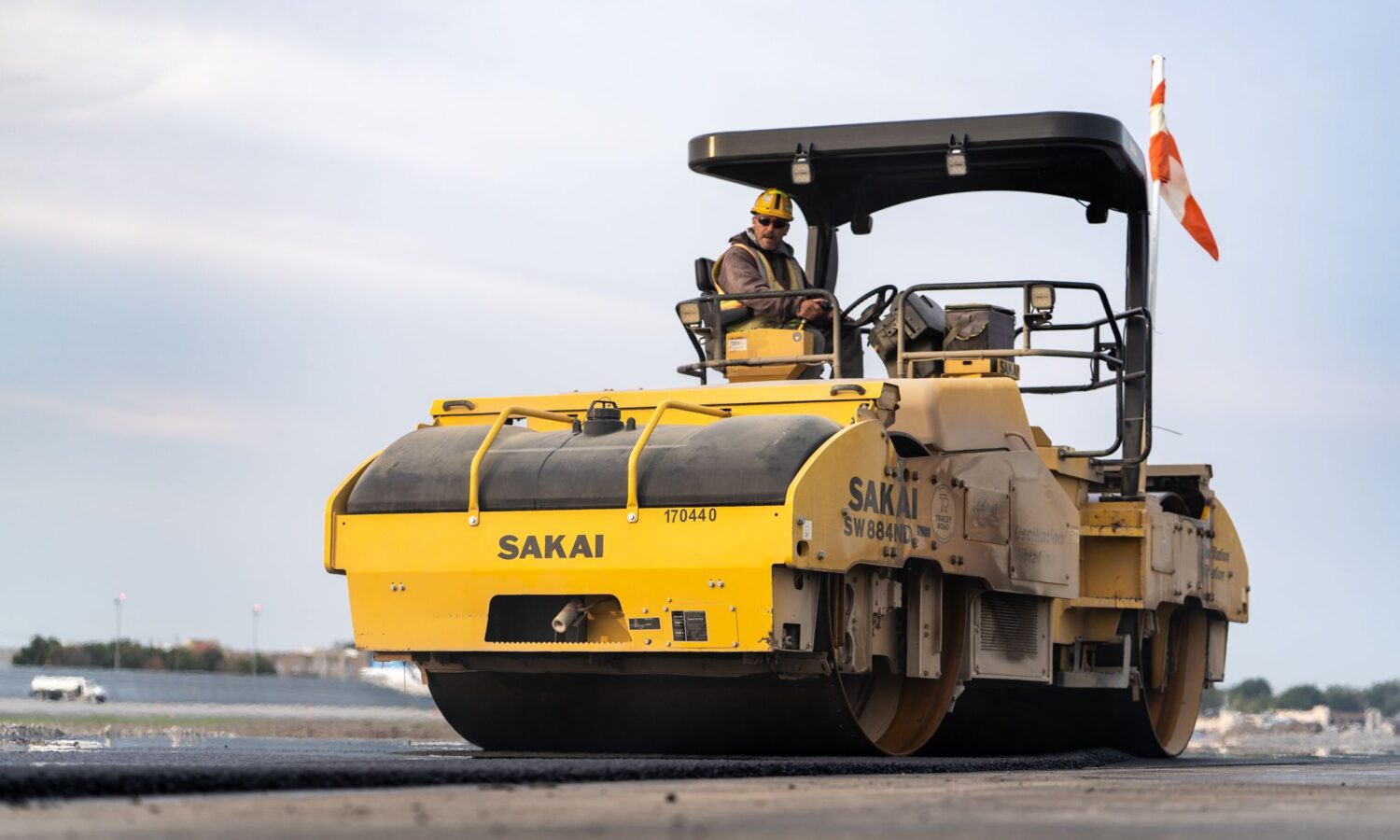 Worker drives yellow road roller, flattening asphalt under grey sky