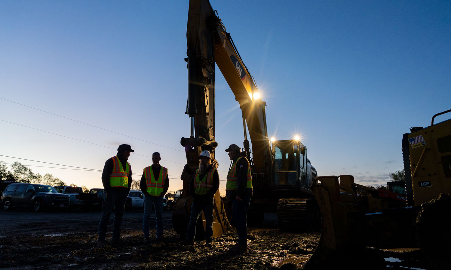 Four workers in vests stand before machinery during night construction, backlit by equipment spotlights