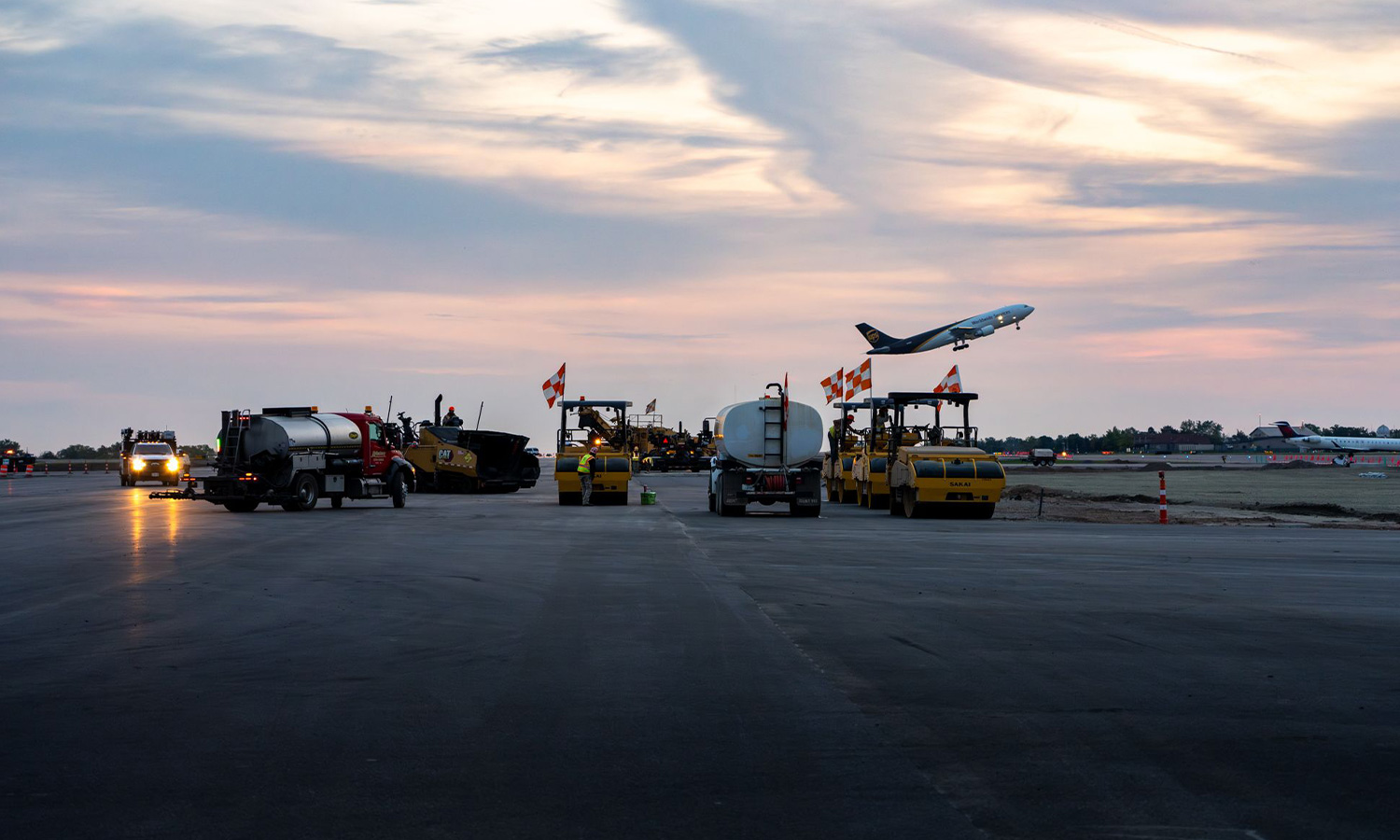 Runway construction at dusk; plane takes off at sunset