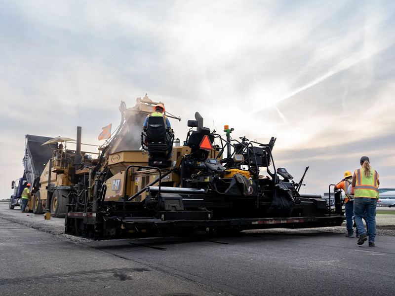 Workers lay new asphalt with heavy machinery under cloudy skies.