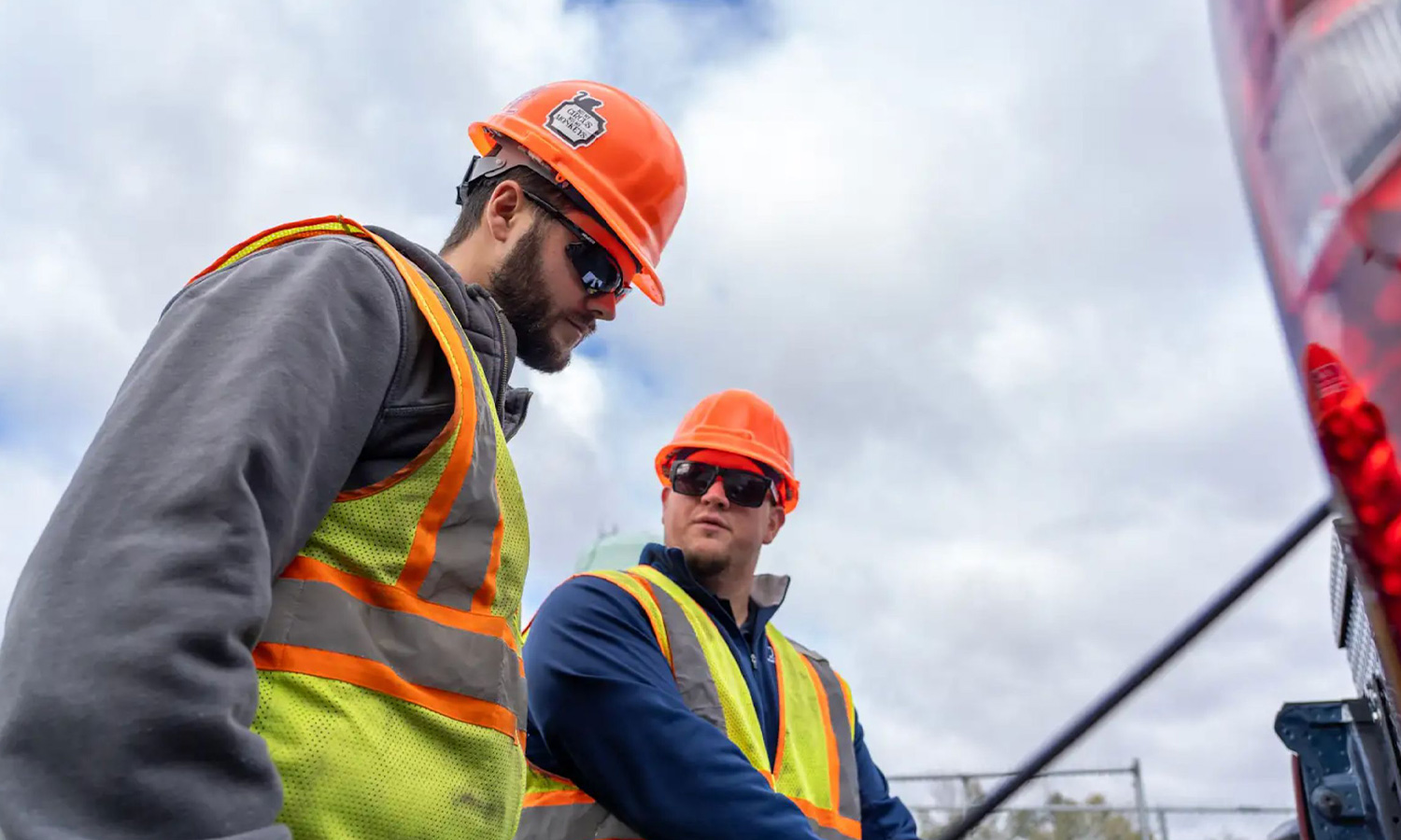 Two workers in safety gear work outdoors under cloudy skies.