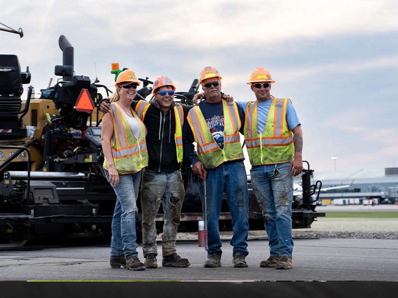 Four construction workers pose before road paving machinery outdoors.