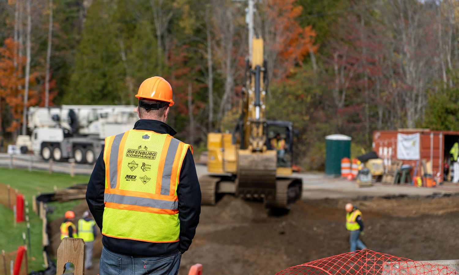 Construction worker watches excavator; others work at site.