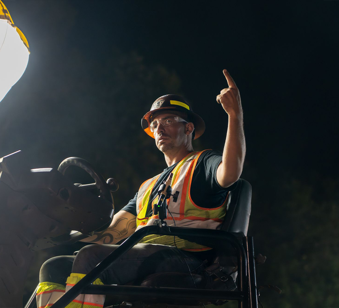 Construction worker gestures upward by work light at night.