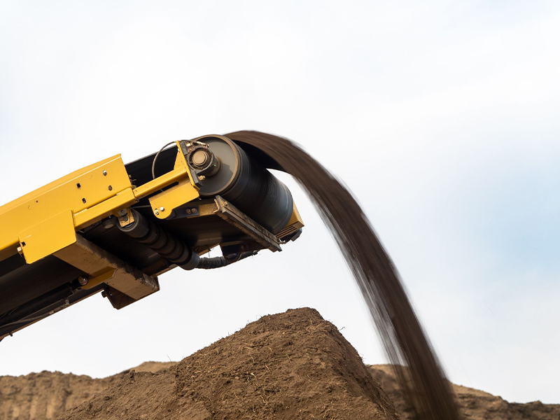 Yellow conveyor deposits soil onto mound beneath cloudy sky.