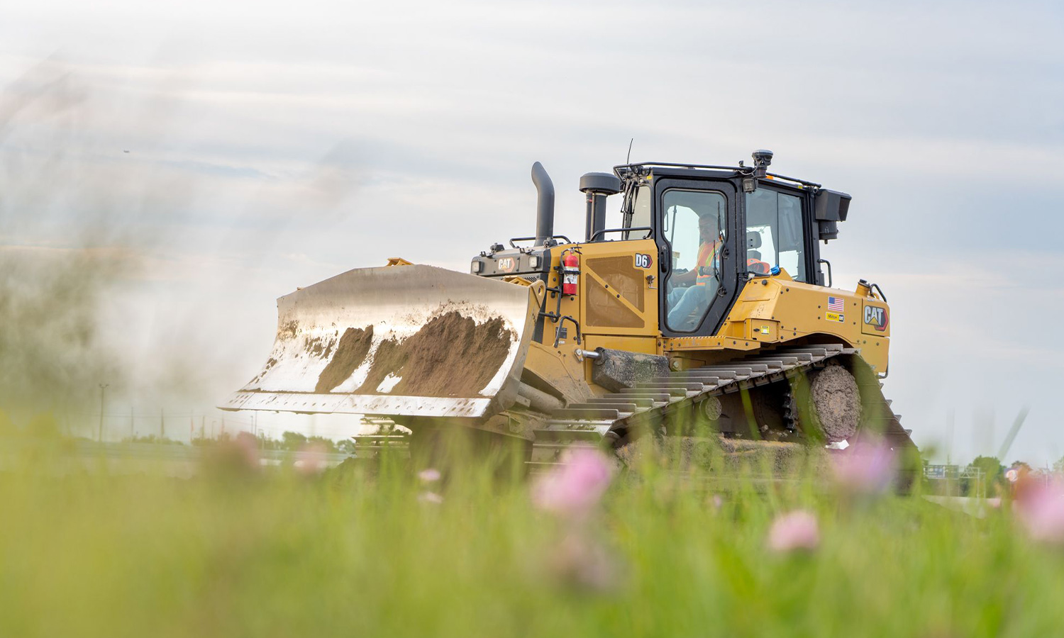 Yellow bulldozer pushes dirt; operator visible, flowers blurred.