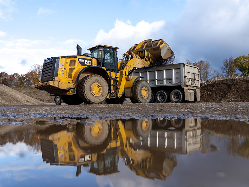 Yellow loader dumps gravel into truck; puddle reflects scene.