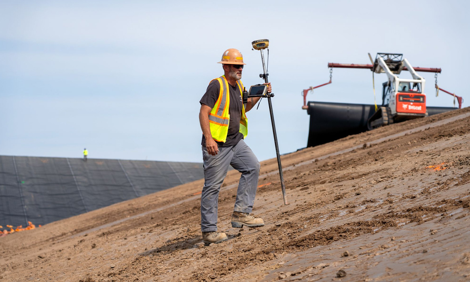 Worker in vest surveys dirt slope; machinery operates behind.