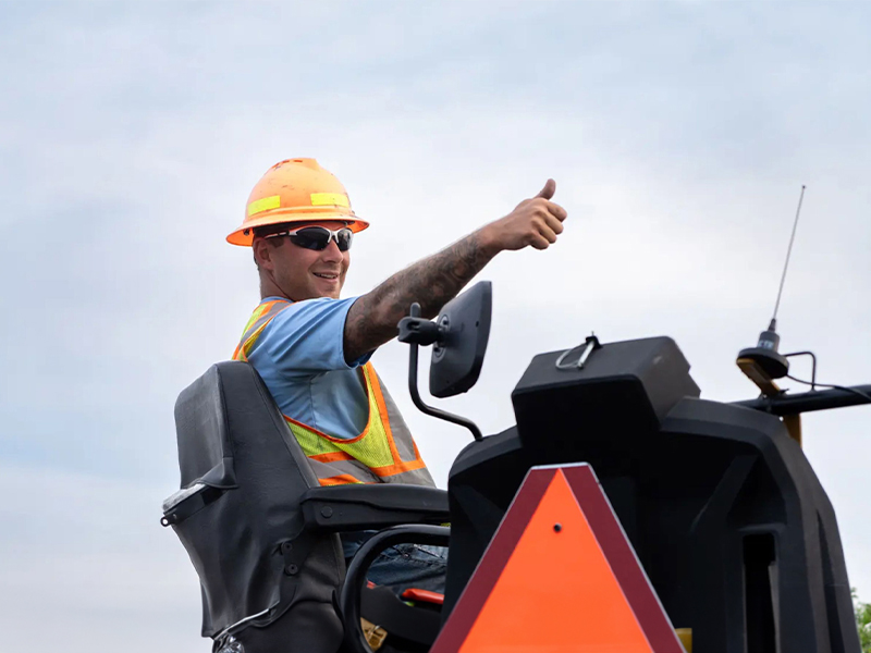 Construction worker in safety gear gives thumbs up on machinery.