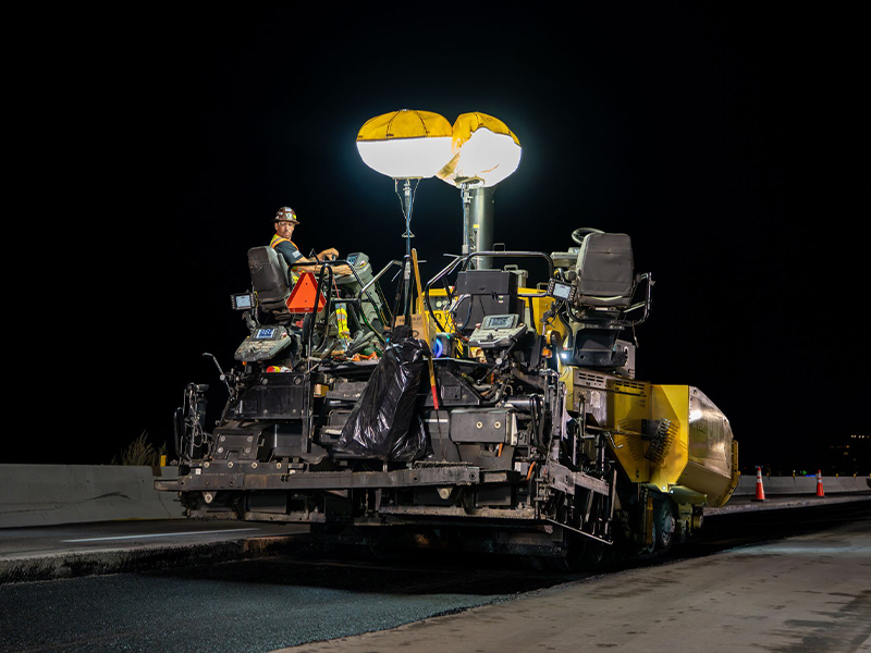 Worker operates asphalt paver under bright night lights.