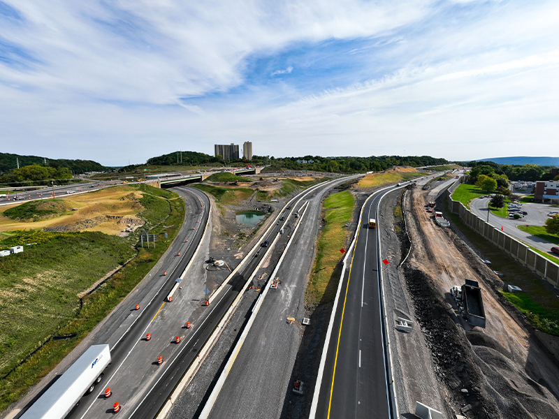 Highway construction: multiple lanes, vehicles, barriers, grassy areas.