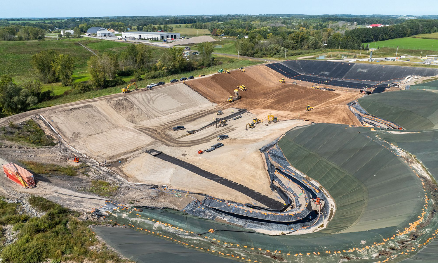 Landfill construction with machines, trucks, liners; fields surround.