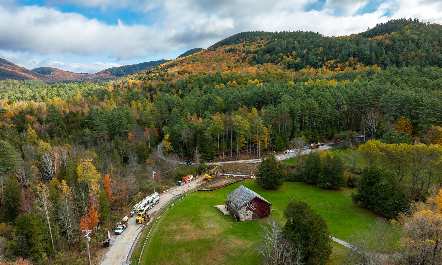 Utility crews work along winding road by red barn, autumn trees.