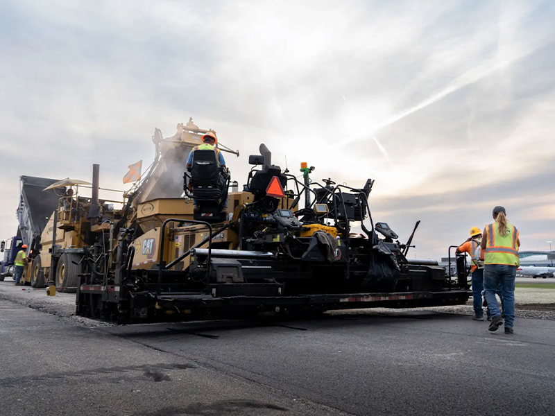 Workers lay new asphalt with heavy machinery under cloudy skies.