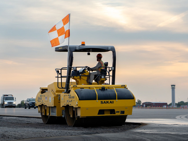 Worker drives yellow Sakai road roller; vehicles, tower behind.
