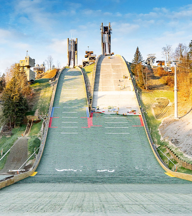 Large ski jump, autumn trees, mountains behind, river in front.
