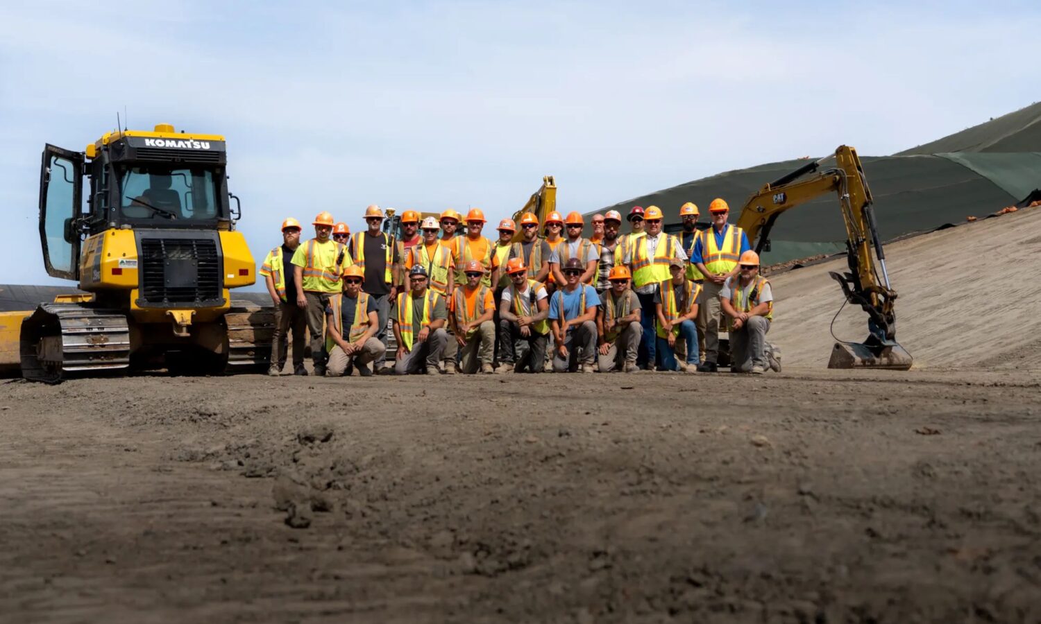 Construction workers in safety gear pose by bulldozer and excavator.