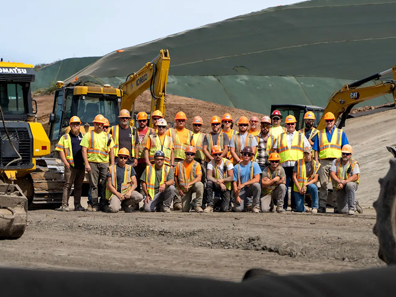 Construction workers in safety gear pose by machinery and slope.